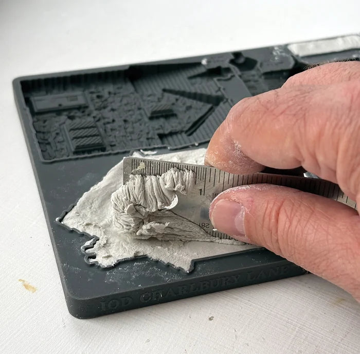 A close-up of a hand using a metal ruler to measure a layer of gray plaster or clay on a black 3D printable terrain mold tray.