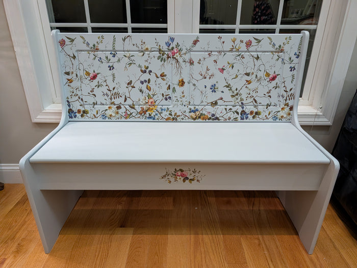 White bench with floral-patterned backrest in a room with wooden floor and window.