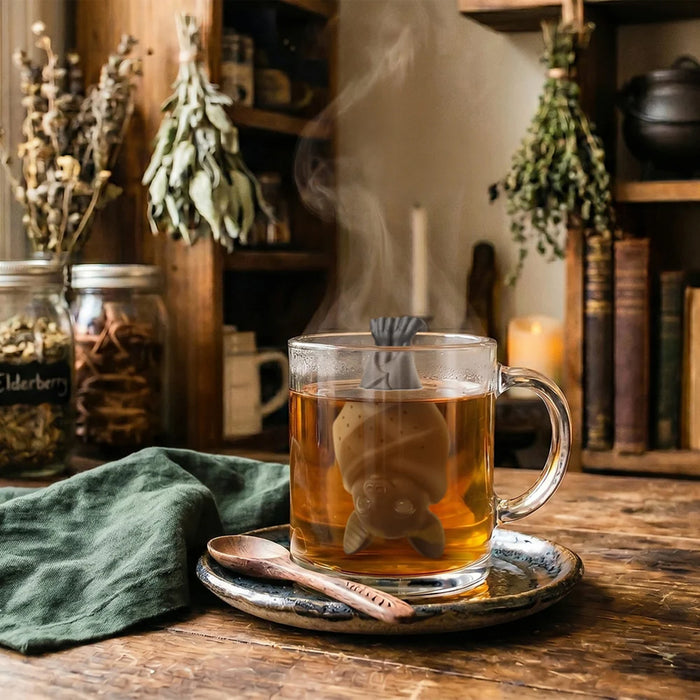 A clear glass mug filled with tea sits on a rustic metal tray with a wooden spoon, set on a wooden table. A bat-shaped tea infuser rests inside the mug, with steam rising.