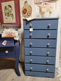 A blue dresser with six drawers, positioned against a wall in a home setting, with a floral pattern on the wallpaper and a framed picture above it.