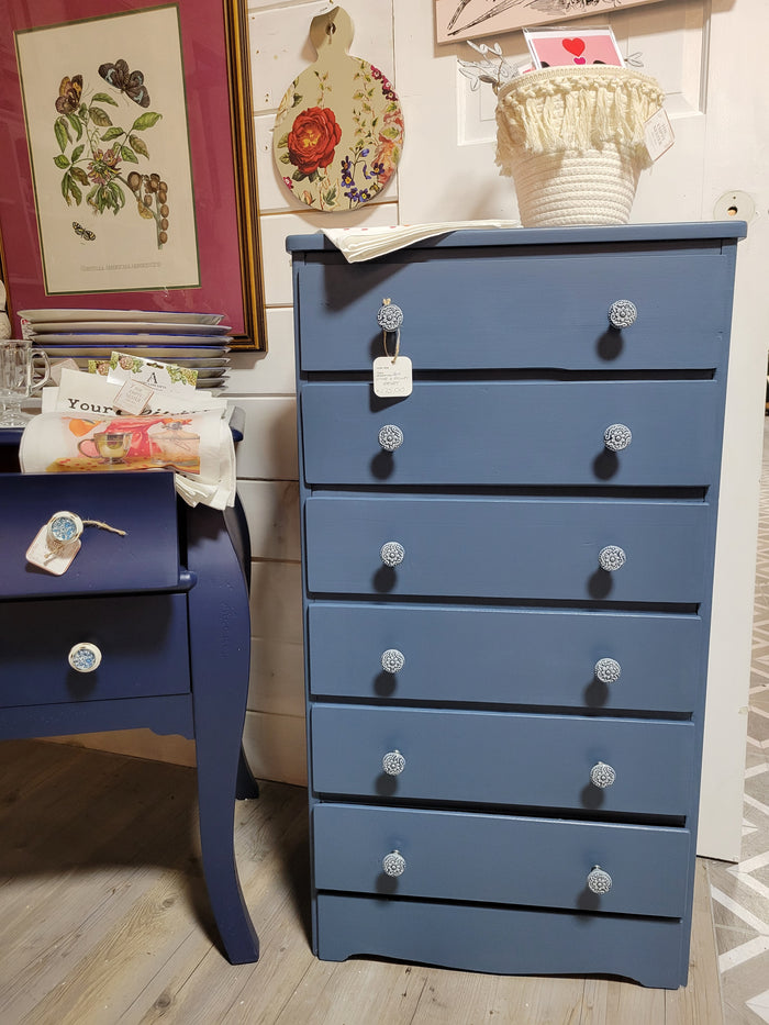 A blue dresser with six drawers, positioned against a wall in a home setting, with a floral pattern on the wallpaper and a framed picture above it.