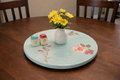 Round wooden tray with floral design on a table with a vase of flowers and small boxes.